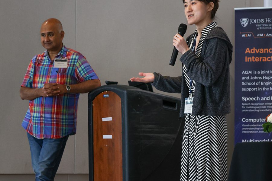 Sanjeev Khudanpur, AI2AI Initiative Director, stands onstage beside Xuan Zhang, 2023-2024 AI2AI Fellow, as she addresses the audience.