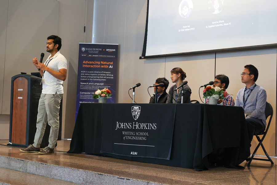 Desh Raj, 2022-2023 AI2AI Fellow, stands onstage and speaks of his AI2AI Fellowship experience while 2023-2024 AI2AI Fellows Ambar Pal, Xuan Zhang, Aniket Roy, and Jiang Liu listen while seated onstage.