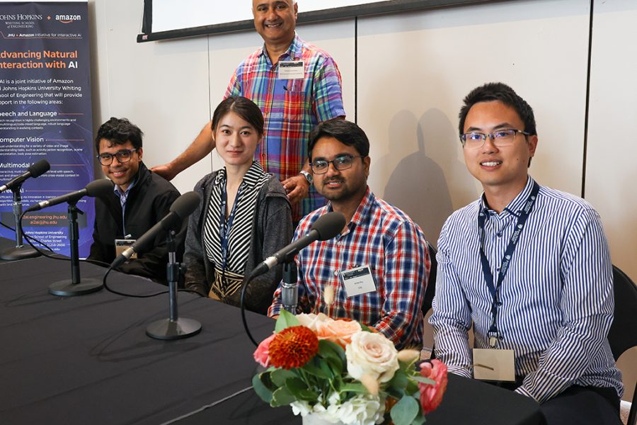 2023-2023 AI2AI Fellows Ambar Pal, Xuan Zhang, Aniket Roy, and Jiang Liu sit behind a table onstage and pose for a photo with Sanjeev Khudanpur, AI2AI Initiative Director.