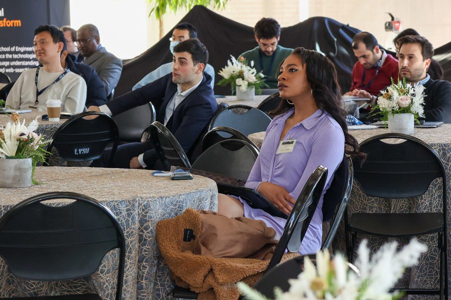 Event attendees sit at round tables and observe the presentation that is being delivered.