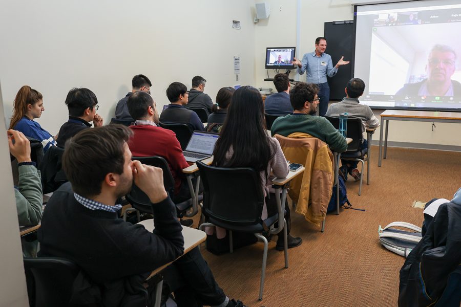 A group of individuals sit in a classroom attending the breakout session focused on general discussion of speech, language, and data-related abstracts. Philipp Koehn, Department of Computer Science, JHU, stands in the front of the room beside a projector screen depicting virtual participants.